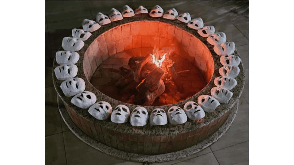 Há um círculo de máscaras de gesso em volta do fogo. Todas brancas, todas vazias e voltadas para o centro, congeladas no instante que antecede a escolha. Nenhuma queima ainda, mas cada uma delas representa uma hora do dia vivida no mais profundo exílio de si: a dedicação incansável, o profissionalismo impecável, a postura que não dá trabalho, a parceria que tudo aguenta e a disponibilidade constante. São rostos moldados para caber nas expectativas do mundo, dispostos em um espaço sagrado e improvisado, onde a imagem fotográfica atua como um espelho e um chamado. Esta fogueira central é o Self convocando à transformação alquímica e lançando um ultimato silencioso: o sacrifício do que se deixará de ser ou a continuidade de uma existência morna e artificial.